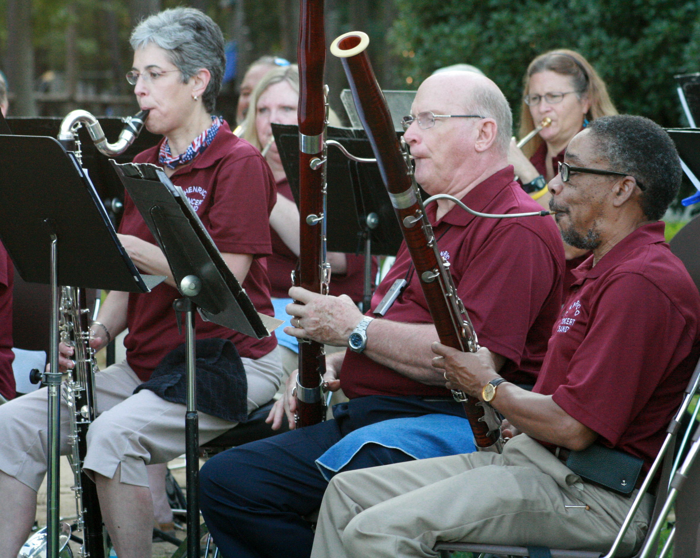 A photo of several concert band members, playing brass and wind instruments in the outdoors.