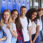 Diverse group of seven smiling teens standing next to blue lockers teens