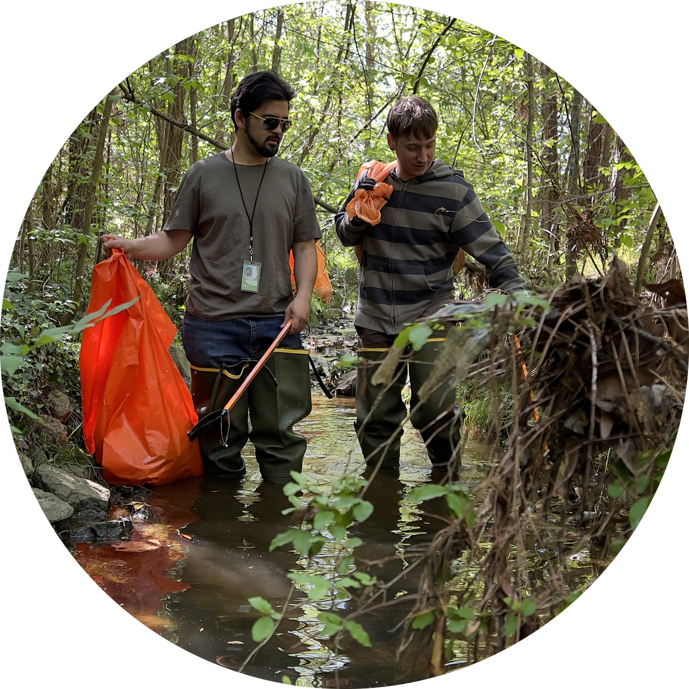HEART water resources circle photo of 2 men wearing knee high rubber boots walking in a stream each one holding an orange trash collection bag & tool for picking up trash