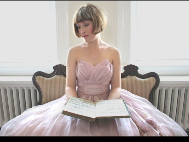 Sarah Walston sits in a pink ball gown with a book on her lap, looking serenely to the left.